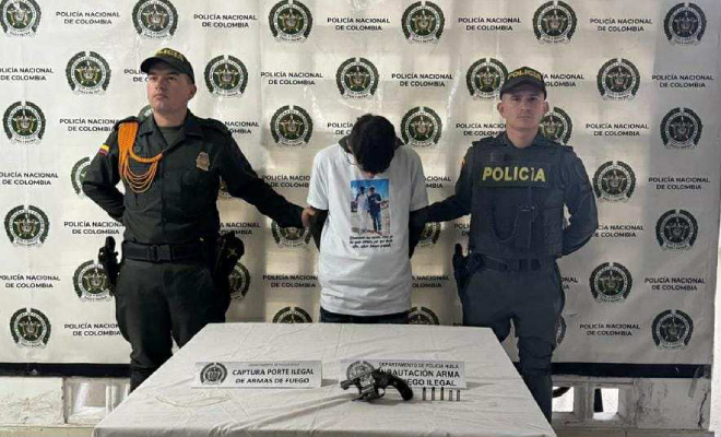 Dos policias con un hombre capturado en el medio, y una mesa con un arma de fuego incautada