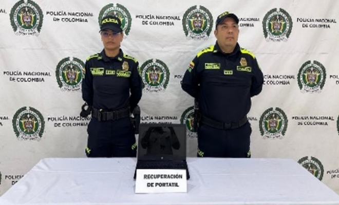 Dos funcionarios policiales frente a pendón institucional, frente a ellos una mesa con computador recuperado
