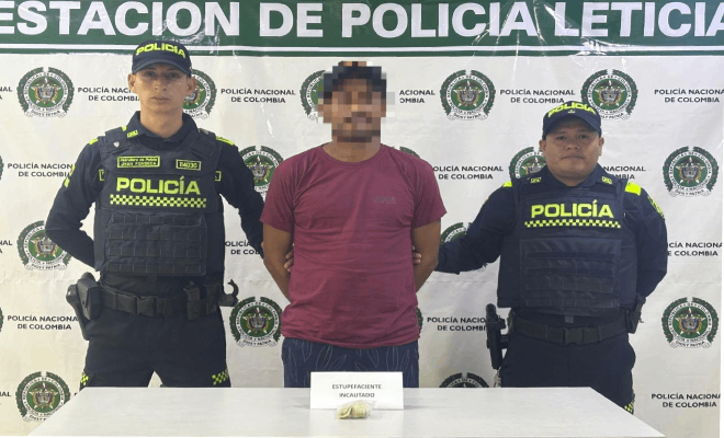 Dos policías y una persona civil se encuentran de pie detrás de una mesa sobre la cual se exhiben estupefacientes incautados, con un banner institucional al fondo