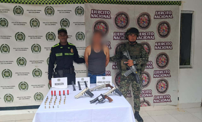 Hombre capturado, al lado un funcionario de la policía y al otro del ejército con una mesa en frente con armas de fuego y municiones.