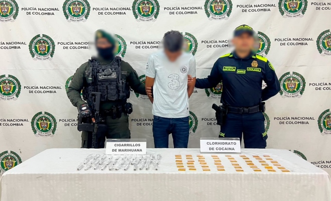 Dos funcionarios policiales con un ciudadano capturado frente a pendón institucional y delante una mesa, sobre la cual esta en material incautado