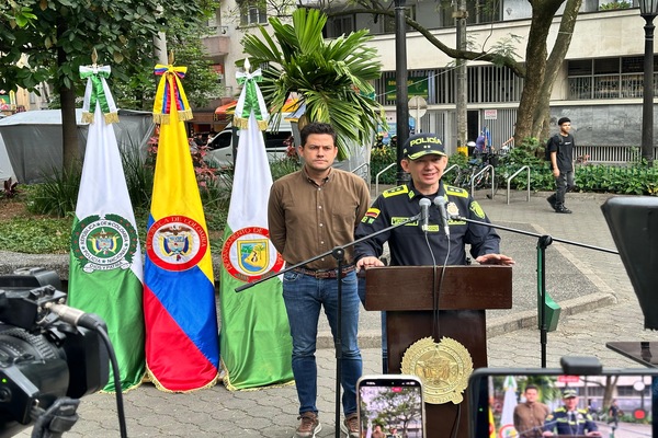 Via pública, tres banderas en hasta, Policía Nacional, Colombia, Medellín frente a ellas, dos personas, un policía y un ciudadano, las cámaras de video en rueda de prensa.   