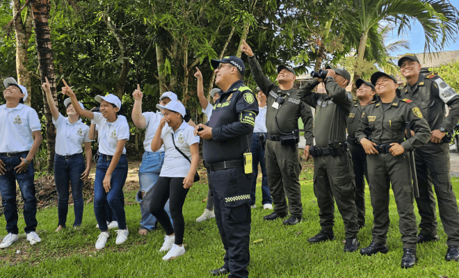 Seis policías de pie observan hacia arriba utilizando binoculares junto a personas de civil con camisas blancas que también miran hacia arriba y señalan