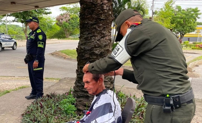 Auxiliar de policía cortando cabello de un ciudadano con otro funcionario policial al fondo