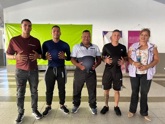 Se observa a cinco personas formando un grupo reducido, posan de forma unida y sonriente, colocando ambas manos sobre su abdomen mientras participan en una dinámica conjunta dentro de un espacio adecuado para actividades formativas y de concientización