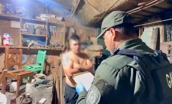 Interior de un taller con herramientas y estanter&iacute;as; una persona uniformada observa un documento mientras otra persona se encuentra de pie frente a ella, y objetos de trabajo est&aacute;n distribuidos sobre mesas y paredes.