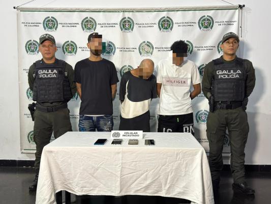 Tres personas con los rostros pixelados permanecen de pie frente a un fondo institucional de la Policía Nacional, acompañadas por dos integrantes del GAULA. Sobre una mesa se exhiben varios teléfonos celulares incautados.