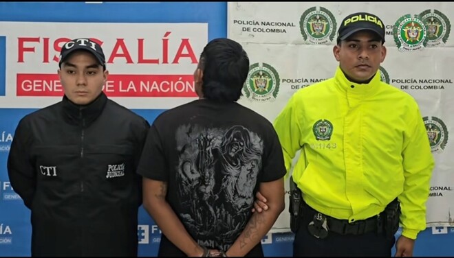 Un hombre esposado de espaldas, junto a un agente del CTI y un oficial de la Policía Nacional, ante el letrero de la Fiscalía General de la Nación