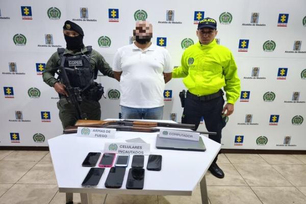 En un espacio institucional, una persona con el rostro difuminado se encuentra de pie entre dos integrantes de la Policía Nacional. Frente a ellos hay una mesa con armas de fuego, varios teléfonos celulares incautados y un computador portátil, debidamente rotulados. Al fondo se observa un respaldo con logos de la Fiscalía, la Alcaldía de Medellín y la Policía Nacional