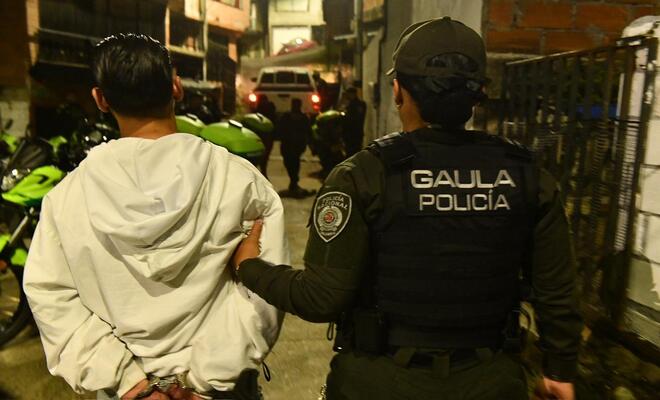 Calle estrecha en zona urbana durante la noche; una persona con chaqueta clara camina con las manos sujetas detrás mientras una persona uniformada con chaleco que dice “GAULA Policía” la guía, con varias motocicletas y un vehículo al fondo