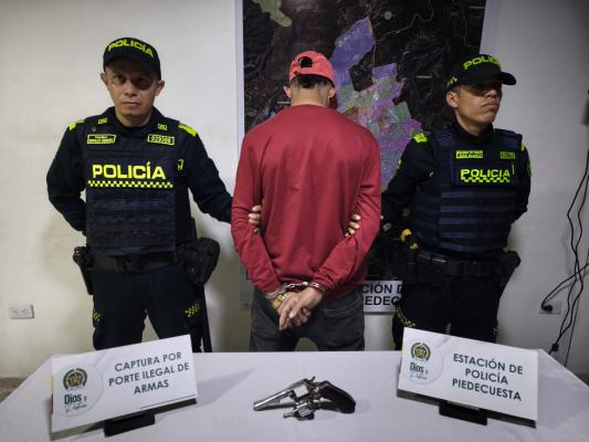 Hombre capturado con buso y gorra roja en medio de dos Polic&iacute;as, en frente sobre una mesa se encuentra el arma de fuego incautada