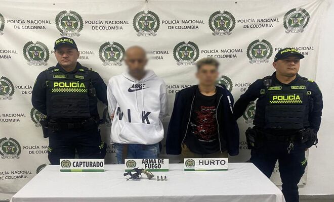En un espacio institucional, dos personas con los rostros difuminados se encuentran de pie entre dos integrantes de la Policía Nacional. Frente a ellos hay una mesa con un arma de fuego, munición y rótulos que indican "capturado", "arma de fuego" y "hurto". 