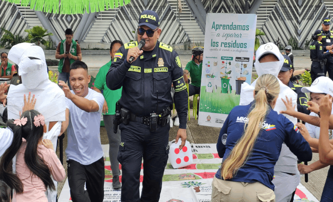 Policía hablando por un micrófono al lado de una funcionaria de migración mientras varios niños y niñas con camisas blancas sonríen
