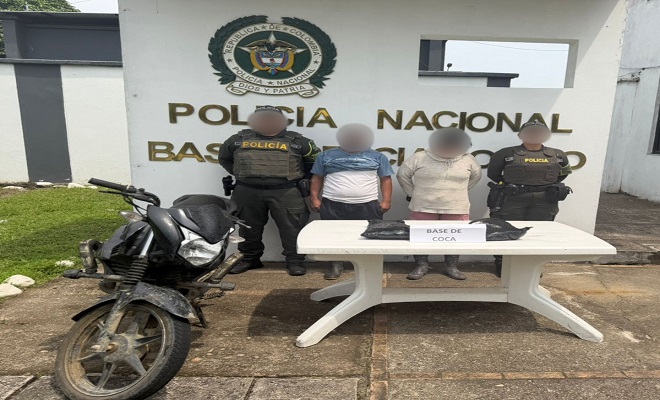 Dos policías junto a dos personas capturadas, en frente una mesa con estupefacientes como evidencia y una motocicleta incautada 