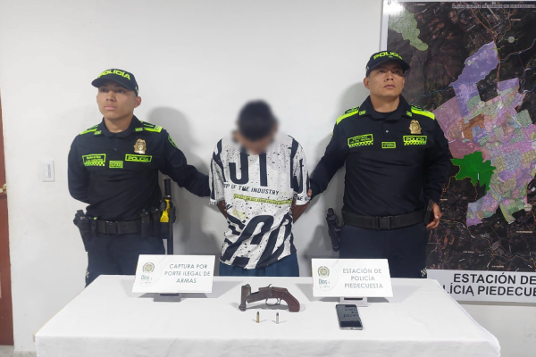 Hombre capturado en medio de dos policías, en frente mesa con arma de fuego incautada