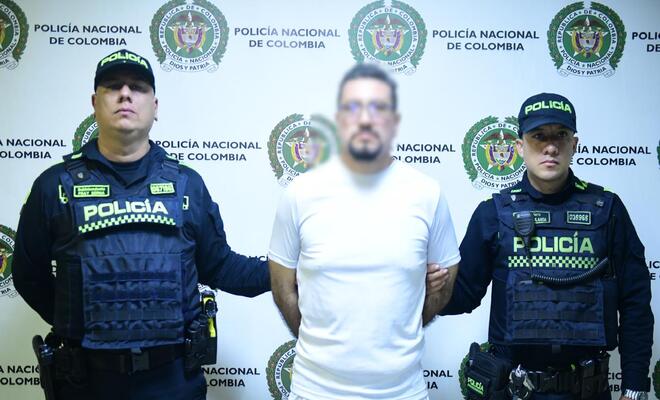 Tres personas de pie frente a un panel institucional de la Policía Nacional de Colombia; la persona ubicada en el centro viste una camiseta blanca y está acompañada a ambos lados por dos personas uniformadas