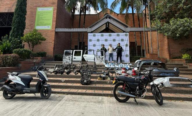 Escalinata exterior frente a un edificio institucional con un fondo de la Polic&iacute;a Nacional de Colombia; en primer plano hay motocicletas, una camioneta y m&uacute;ltiples autopartes organizadas sobre los escalones, mientras varias personas se encuentran de pie detr&aacute;s.