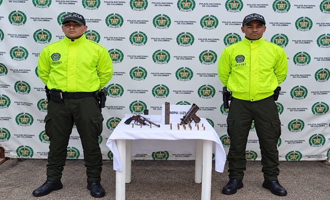 Dos uniformados de la Policía Nacional custodian una mesa donde se observan dos armas de fuego artesanales y varios cartuchos de munición incautados durante un procedimiento realizado en el departamento del Chocó