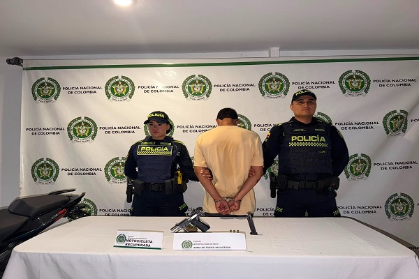 Dos policías custodian a un hombre capturado junto a una mesa con un arma de fuego incautada y una motocicleta recuperada