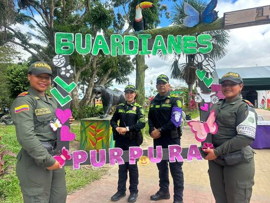  Cuatro funcionarios posan frente al mismo arco decorado; se aprecian detalles de sus uniformes y elementos alusivos a la campaña, con un entorno natural y esculturas al fondo