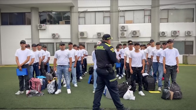 Se desarrolla un acto de recibimiento en un espacio abierto frente a una construcción, donde un oficial de policía se sitúa al frente de una fila de jóvenes con camisetas blancas. Se aprecian pertenencias personales en el suelo, lo que indica que se está dando la bienvenida a un grupo de personas que se inician en un proceso relacionado con la institución policial